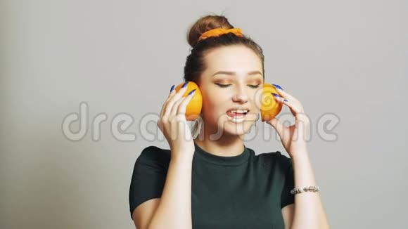 漂亮的女孩把橘子的一半水果放进耳朵里就像耳机一样青少年在演播室背景下玩得很开心概念时尚视频的预览图