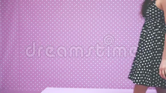 一个身材瘦削的黑发女孩头上戴着皮钉坐在粉红色的长凳上享受着她漂亮的波尔卡点裙视频的预览图