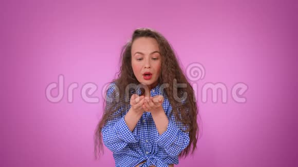 一个漂亮的卷发女孩从手掌上吹出纸屑笑容满面一个女孩在一个孤立的粉红色背景上的肖像视频的预览图