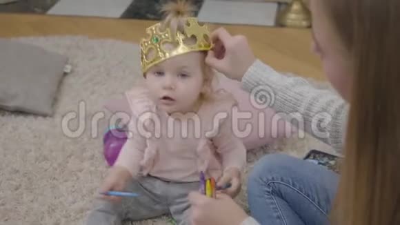 一位年轻的白人母亲试图给女儿戴上玩具王冠的特写镜头漂亮的金发女婴坐在柔软的视频的预览图