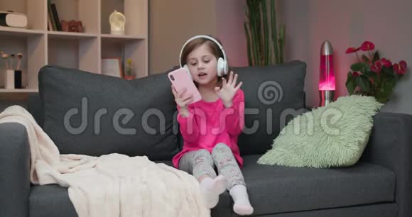 戴着耳机的小女孩在沙发上跳舞用智能手机触摸屏幕现代科技和快乐视频的预览图