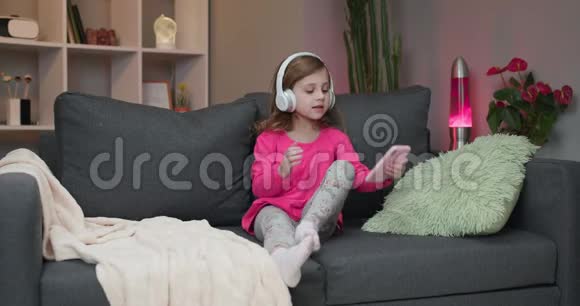 可爱的小白种人女孩坐在家里的沙发上微笑着用现代智能手机耳机听音乐视频的预览图