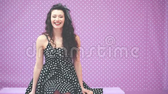 一个穿着漂亮裙子的金发女孩坐在粉红色的长凳上微笑着看着相机很可爱视频的预览图