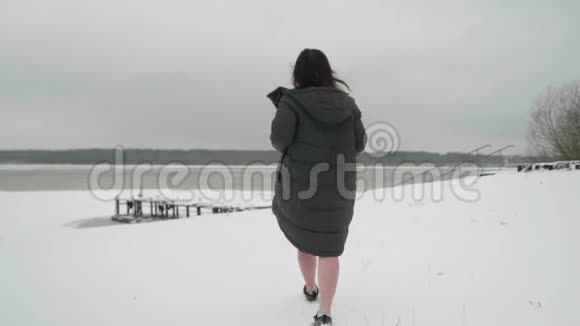 在雪地上行走的女人的后景把她的外套脱掉向前移动到覆盖着冰雪的池塘慢动作视频的预览图