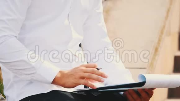 一个年轻的面目全非的商人穿着白衬衫带着剪贴板和笔坐在外面的城市里的长凳上视频的预览图
