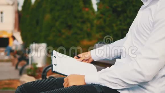 一个年轻的面目全非的商人穿着白衬衫带着剪贴板和笔坐在外面的城市里的长凳上视频的预览图