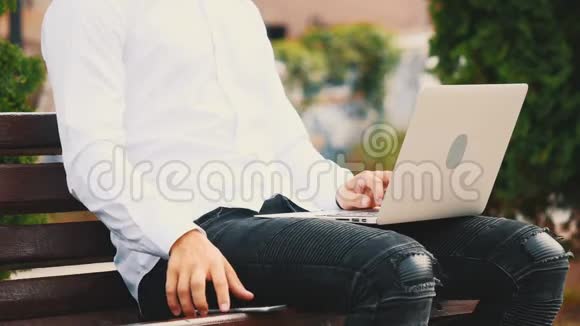 一个年轻的不可辨认的商人正在笔记本电脑上工作穿着白色衬衫坐在城外的长凳上作物视频的预览图