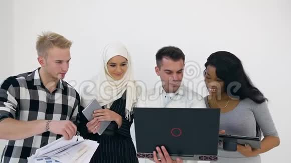 四个年轻的多种族有目的的办公室工作人员使用笔记本电脑和在白色墙壁背景上的杜斯克联合项目视频的预览图