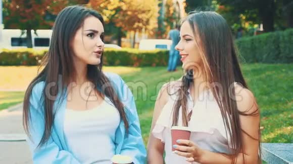 快乐的女孩朋友们正在谈论和微笑手里拿着咖啡坐在公园的长凳上行动行动4K视频的预览图