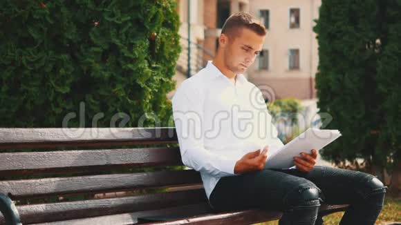 一位年轻的商人正在翻阅重要的文件穿着白衬衫坐在城外的长凳上视频的预览图