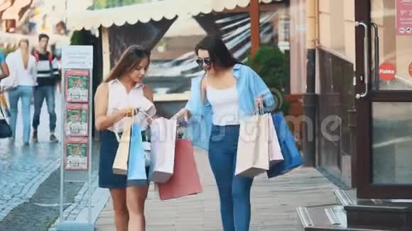 两个有魅力的年轻女人正拿着购物袋在夏天的街道上散步和交谈快关门复制空间4K视频的预览图