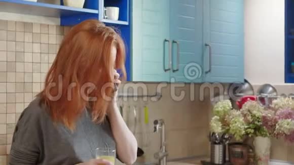 迷人的年轻红发女孩周末在厨房喝橙汁使用智能手机与父母交谈视频的预览图