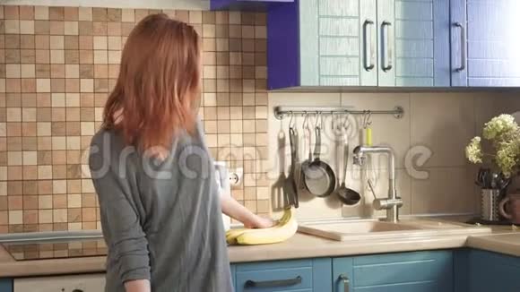 迷人的饥饿红发女孩在周末的清晨在厨房里吃一根大香蕉把皮洗干净视频的预览图