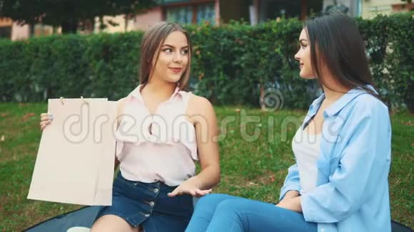 两个漂亮的女孩坐在公园的长凳上金发女孩给她的黑发女人买了一件新的脱光上衣视频的预览图