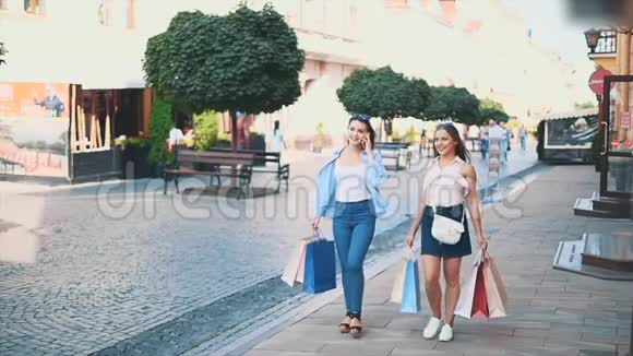 两个有魅力的年轻女人正拿着购物袋在夏天的街道上散步和交谈其中一个在打电话视频的预览图