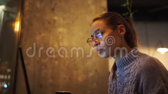 年轻女性自由职业者晚上坐在咖啡馆里用笔记本电脑看监视器视频的预览图
