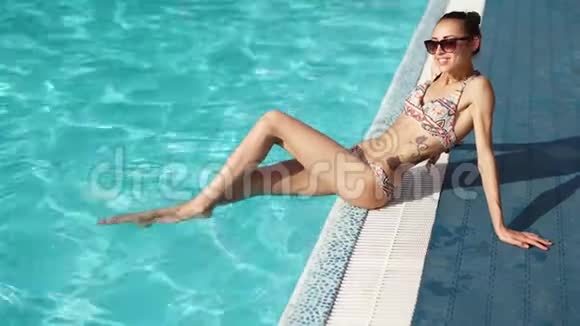 年轻性感晒黑比基尼女人坐在池边双腿在水中享受阳光明媚的一天视频的预览图