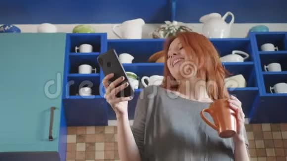 跳舞乐趣在智能手机上听音乐漂亮的红发女孩在家庭厨房喝早咖啡视频的预览图