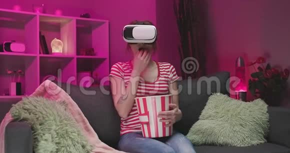 年轻女子戴VR耳机晚上用爆米花看电影坐在沙发上的女人戴着VR眼镜视频的预览图