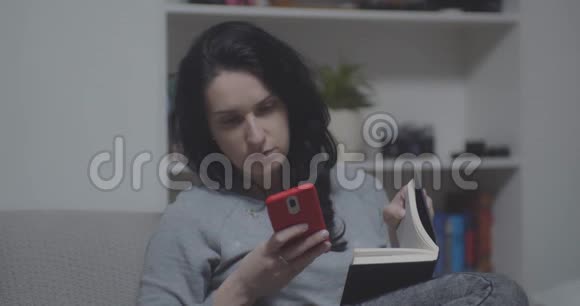 年轻女性在家沙发上看书使用智能手机视频的预览图