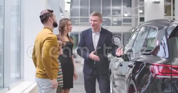 自信的男性汽车经销商在汽车陈列室向高加索家庭宣传新车并摇动的年轻人视频的预览图
