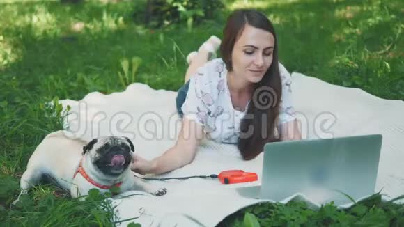 女孩躺在公园草坪上的地毯上她在笔记本电脑上打字帕格躺在旁边环顾四周视频的预览图