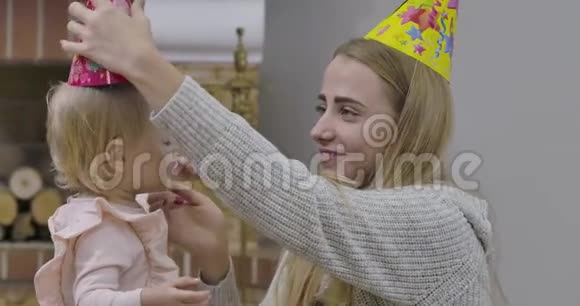 年轻的高加索女人在小女孩头上戴派对帽穿着粉色上衣庆祝她第一个生日的漂亮宝宝视频的预览图