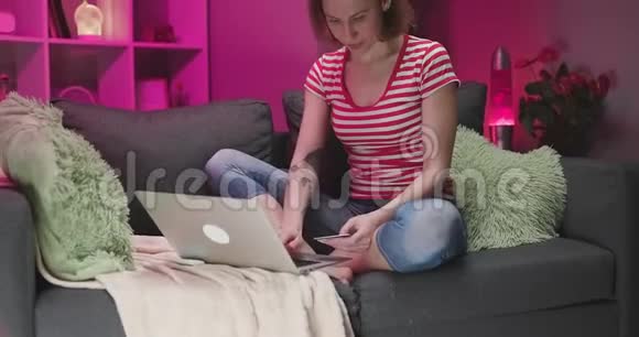 年轻女子坐在沙发上用信用卡在线购买女人用笔记本电脑上网视频的预览图