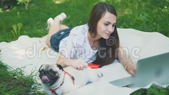 女孩躺在公园草坪上的地毯上她在笔记本电脑上打字帕格躺在旁边环顾四周视频的预览图