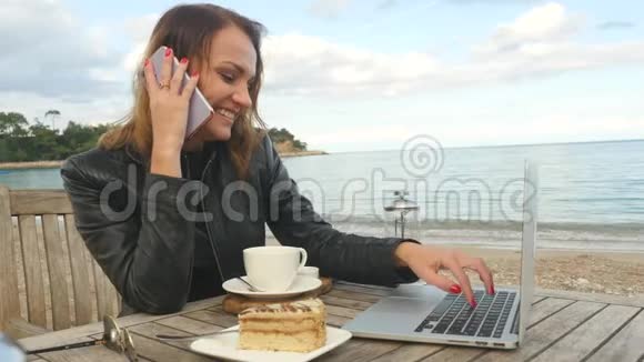 在户外咖啡馆里一个开放的笔记本电脑前年轻的女人在手机上说话视频的预览图