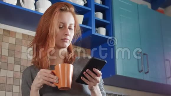 漂亮的红发女孩在家厨房喝早咖啡自由职业者的早晨使用智能手机手表视频的预览图
