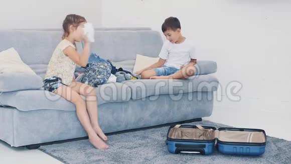 小孩子在家里把衣服扔进手提箱他们坐在蓝色沙发上特写4K视频的预览图