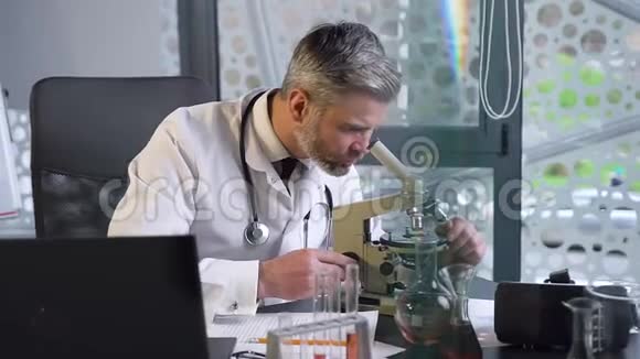 在临床实验室工作的迷人的受人尊敬的白发男医生的正面视角看显微镜和视频的预览图