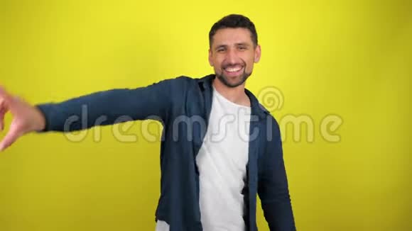 一个穿着白色T恤和蓝色衬衫的年轻人正在黄色背景上带着复制空间微笑着跳舞视频的预览图