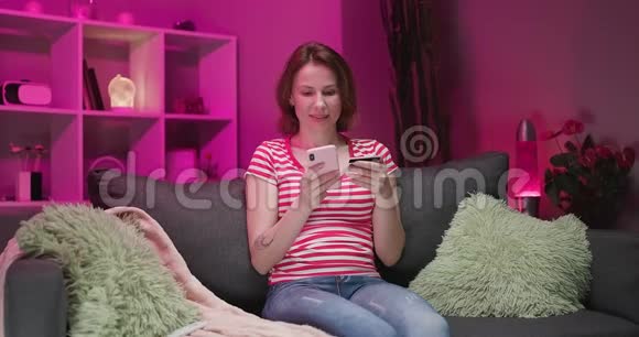 女人在家里坐在沙发上用信用卡和微笑在线购物女人用的是智能手机视频的预览图