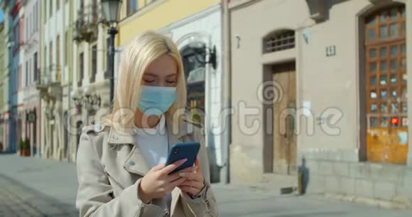 年轻女子戴着防护面具在老城区的街道上使用智能手机担心的女孩搜索关于Covid19的新闻视频的预览图