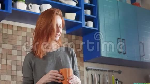 漂亮的红发女孩在家厨房喝早咖啡在智能手机上说话早上打个电话视频的预览图