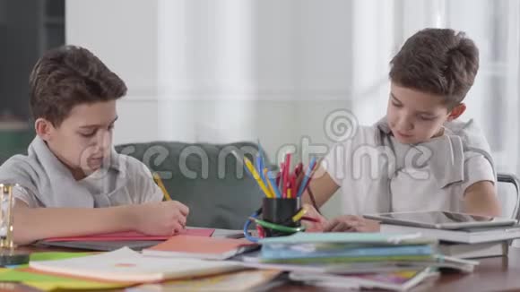 白人学童坐在桌子旁做作业的肖像布鲁内特男孩看着他孪生兄弟视频的预览图