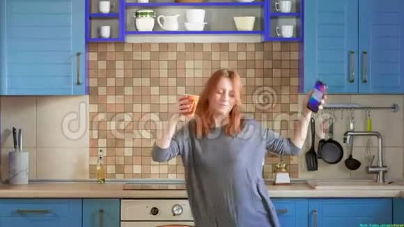 跳舞乐趣在智能手机上听音乐漂亮的红发女孩在家庭厨房喝早咖啡视频的预览图