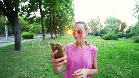 年轻开朗微笑的女人站在夕阳城市绿色公园的背景上用手机自拍视频的预览图
