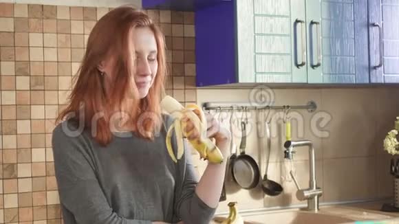 迷人的饥饿红发女孩在周末的清晨在厨房里吃一根大香蕉把皮洗干净视频的预览图