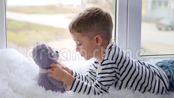 一个可爱的小男孩与泰迪熊玩具分享秘密而躺在窗台上的阴天儿童梦想和视频的预览图
