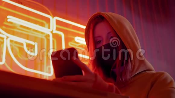 年轻漂亮的时尚女孩用黑色医疗面具的智能手机以霓虹灯为背景的酒吧或俱乐部视频的预览图