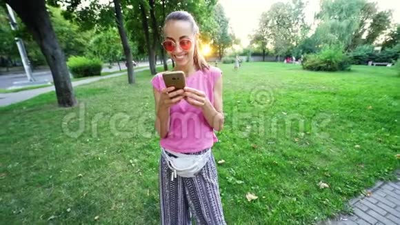 戴着粉红色眼镜的微笑女人站在夕阳城市绿色公园的背景上用手机视频的预览图
