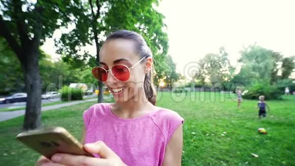 微笑的女人带着滑稽的脸和粉红色的眼镜站在夕阳城市绿色公园的背景上用手机视频的预览图