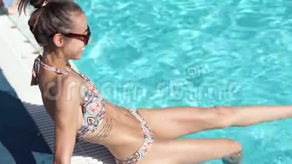 快乐苗条性感微笑比基尼女人戴着黑色太阳镜泼水双腿坐在游泳池旁视频的预览图