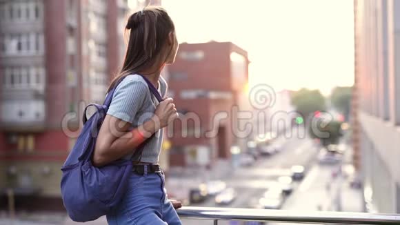 幸福微笑的年轻女子穿着休闲装小背包坐在阳台的栏杆上视频的预览图