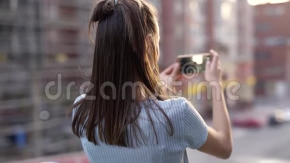 年轻女子站在日落城街道背景下用智能手机拍照或录像视频的预览图