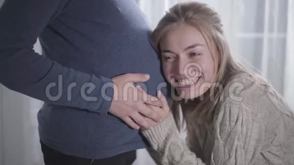 微笑的白种人女人亲吻男性怀孕的腹部和倾听婴儿心跳的肖像有趣的一对视频的预览图