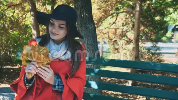 年轻可爱的女孩正拿着一束秋叶一边坐在长凳上背景模糊复制空间4K视频的预览图